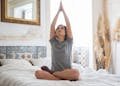A woman begins her day with yoga, sitting on a bed in a serene, well-decorated bedroom.